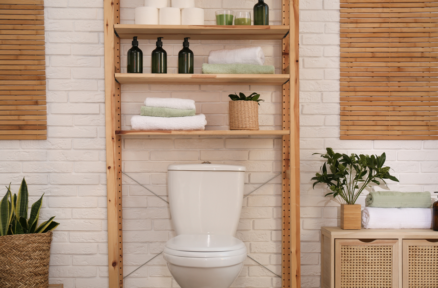 Over-toilet storage shelving unit with folded towels, bottles, and plants in a white bathroom - a practical storage idea for small bathrooms