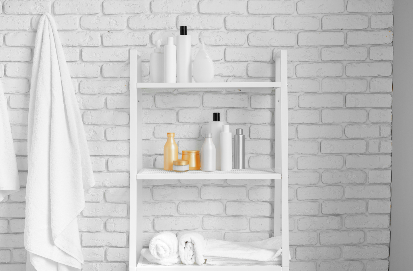 A white metal narrow shelving unit standing against a white brick wall, holding bottles, jars, and rolled towels on multiple shelf levels