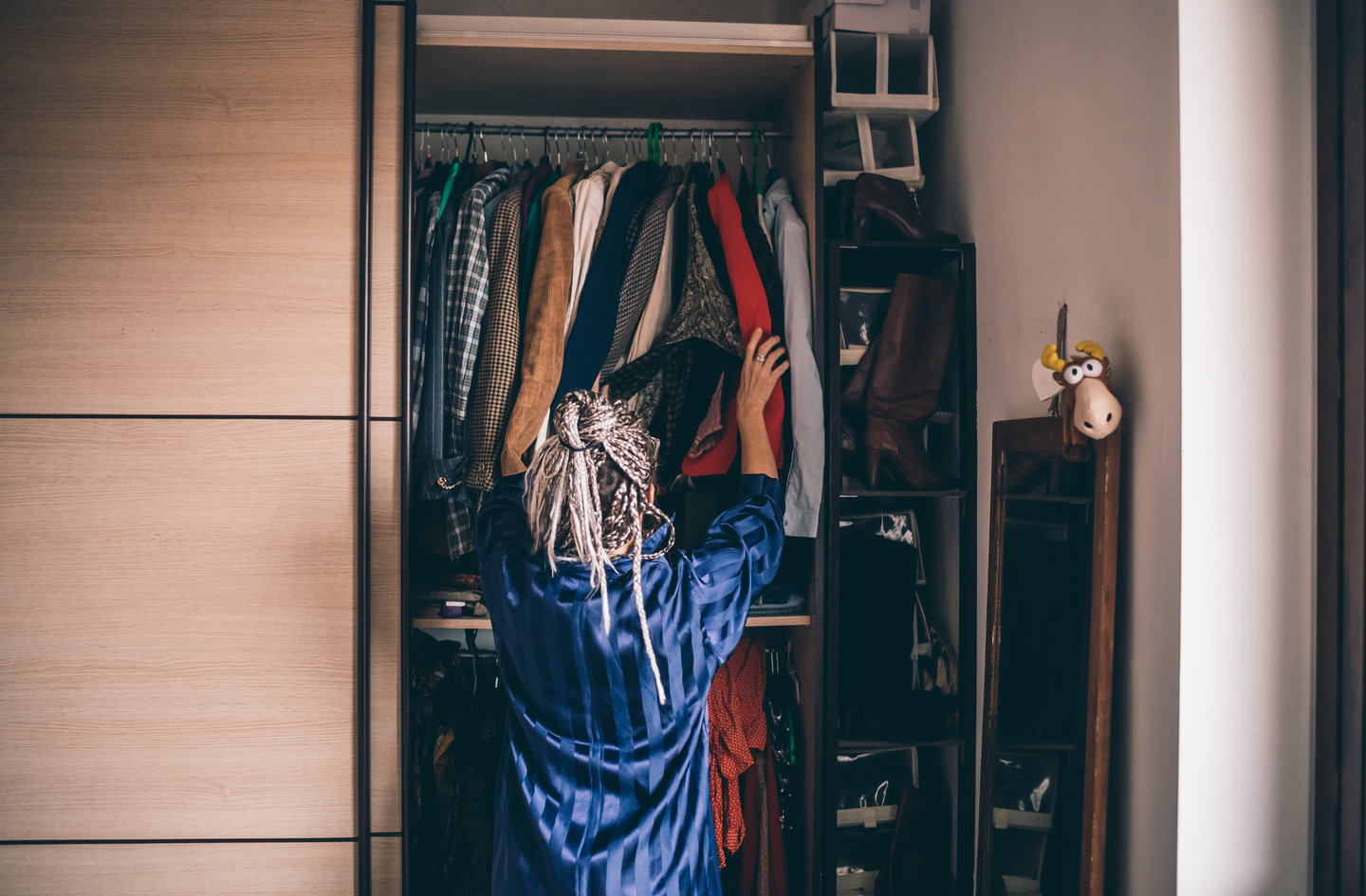 How to organize a small closet with lots of clothes: a person reaches up to pick a garment from a crowded hanging rack while several pairs of boots sit on a shelving unit beside the closet.
