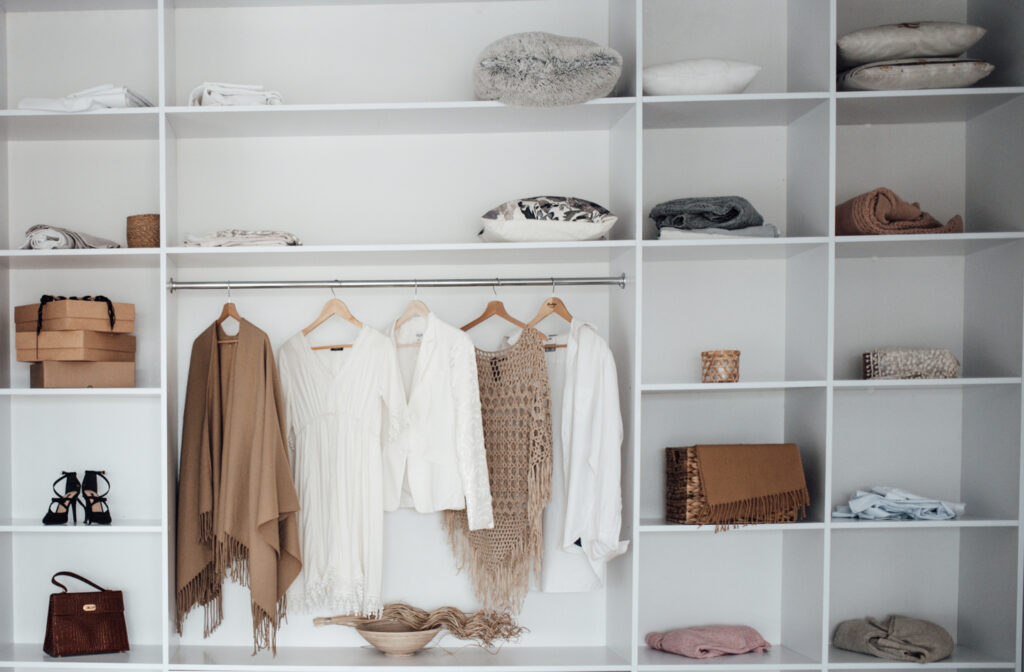 Small closet storage in a white closet with folded linens and boxes on upper shelves, dresses and jackets on a hanging rod, and a handbag and woven basket below