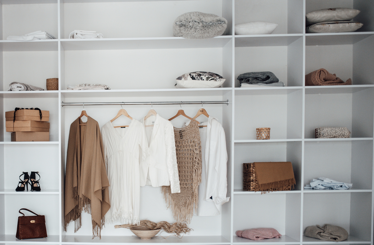 Small closet storage in a white closet with folded linens and boxes on upper shelves, dresses and jackets on a hanging rod, and a handbag and woven basket below