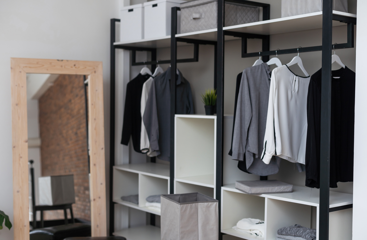 Clothing storage ideas for small bedrooms: an open wardrobe unit holds hanging clothes above folded garments on shelves and in storage boxes, with a tall wood-framed floor mirror standing to its left.